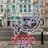 The image features a neon sign with an abstract design depicting elements like a building structure and a fish. The sign includes text that is mirrored, displayed in vibrant colors. It hangs against a backdrop of a street view with classic architectural buildings in the background.