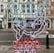 The image features a neon sign with an abstract design depicting elements like a building structure and a fish. The sign includes text that is mirrored, displayed in vibrant colors. It hangs against a backdrop of a street view with classic architectural buildings in the background.