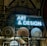 A display features a neon sign with the words 'ART & DESIGN' above shelves containing a variety of plates and pottery. A stained-glass artwork depicting a figure holding a book adds a historical and artistic touch. The background reveals an ornate ceiling adorned with elaborate patterns and designs.