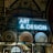 A display features a neon sign with the words 'ART & DESIGN' above shelves containing a variety of plates and pottery. A stained-glass artwork depicting a figure holding a book adds a historical and artistic touch. The background reveals an ornate ceiling adorned with elaborate patterns and designs.